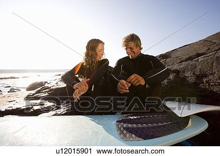 Stock Image - Couple sitting on beach with surfboard smiling.. Fotosearch