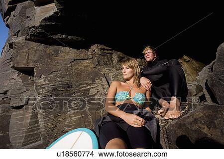 Picture - Couple sitting on large rocks with a surfboard.. Fotosearch