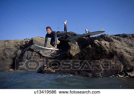 Stock Photograph - Man jumping onto surfboard with other man sitting on rocks behind him.. Fotosearch