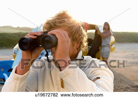 Stock Image - Man on beach with binoculars with woman unloading van at the beach.. Fotosearch