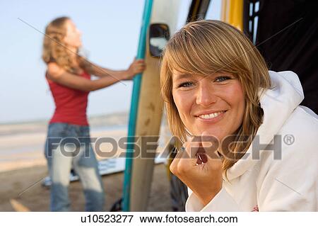 Stock Photo - Woman at the beach smiling with other woman in background holding surfboard.. Fotosearch