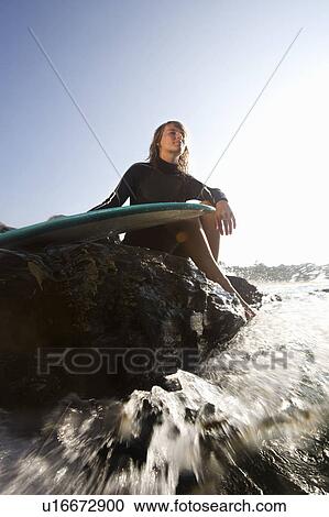Stock Image - Woman sitting on large rocks with surfboard.. Fotosearch