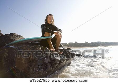 Stock Photography - Woman sitting on large rocks with surfboard smiling.. Fotosearch