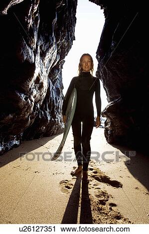 Stock Image - Woman standing with surfboard between large rocks smiling.. Fotosearch