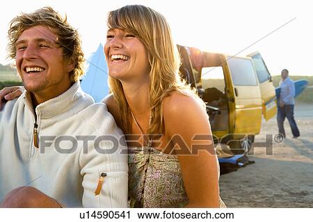 Couple sitting on beach smiling with man carrying surfboard in background. View Large Photo Image Stock Image - Couple sitting on beach smiling with man carrying surfboard in background.. Fotosearch