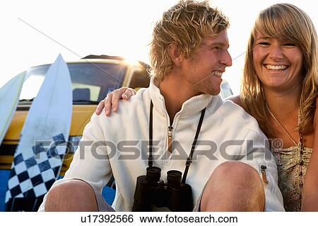 Stock Photograph - Couple sitting on beach smiling with van and surfboards in background.. Fotosearch