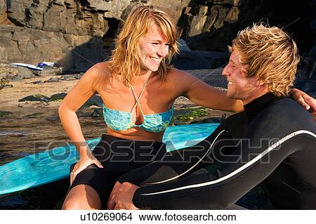 Picture - Couple sitting on large rocks with surfboards laughing.. Fotosearch