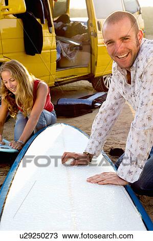 Stock Image - Couple waxing surfboards near a van.. Fotosearch