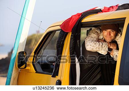 Stock Photograph - Man in van smiling with surfboard leaning against the hood.. Fotosearch