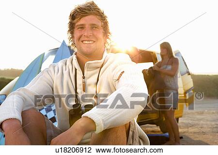 Man sitting on beach smiling with woman unloading van at the beach. View Large Photo Image Stock Image - Man sitting on beach smiling with woman unloading van at the beach.. Fotosearch