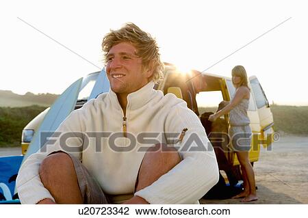 Stock Image - Man sitting on beach smiling with woman unloading van at the beach.. Fotosearch
