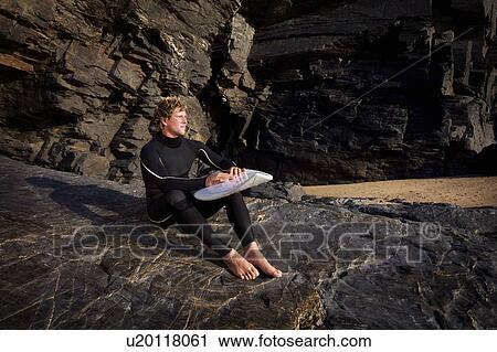 Man sitting on large rock in wetsuit with surfboard on lap. View Large Photo Image Stock Image - Man sitting on large rock in wetsuit with surfboard on lap.. Fotosearch