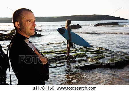 Man standing smiling with woman holding surfboard in background. View Large Photo Image Stock Photograph - Man standing smiling with woman holding surfboard in background.. Fotosearch