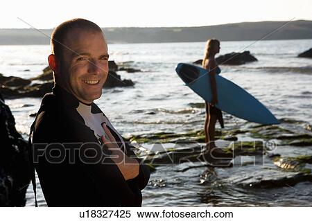 Man standing smiling with woman holding surfboard in background. View Large Photo Image Stock Photography - Man standing smiling with woman holding surfboard in background.. Fotosearch