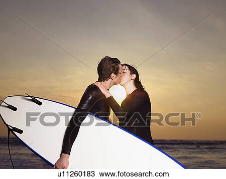 Stock Image - Surfer couple kissing on a beach.. Fotosearch