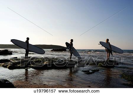 Stock Photo - Three people holding surfboards standing on large rocks.. Fotosearch