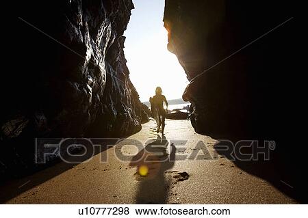 Stock Photo - Woman carrying surfboard through large rocks.. Fotosearch