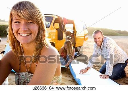 Picture - Woman on the beach smiling with couple waxing surfboards in background.. Fotosearch
