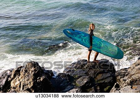 Stock Photo - Woman standing on large rocks with a surfboard smiling.. Fotosearch