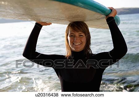 Stock Photo - Woman standing with a surfboard over her head smiling.. Fotosearch