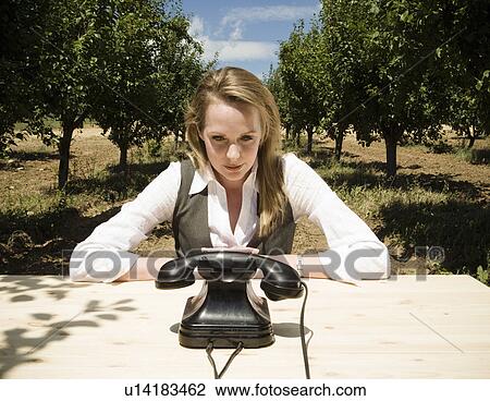 Stock Image - Woman sitting at desk with old telephone in orchard.. Fotosearch