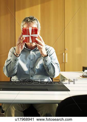 Businessman sitting at desk holding up present View Large Photo Image Stock Image - Businessman sitting at desk holding up present. Fotosearch