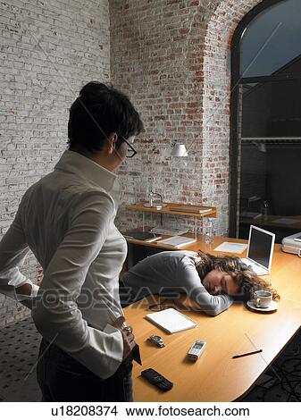 Picture - Businesswoman watching female colleague sleeping on desk. Fotosearch
