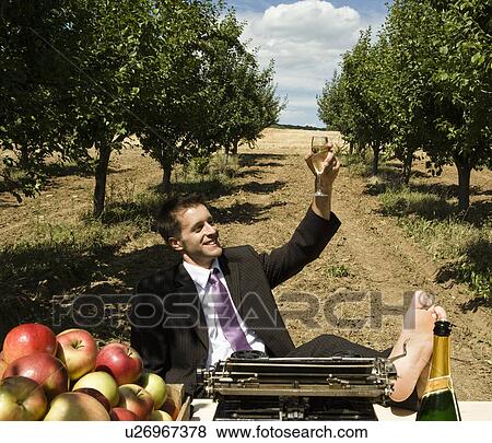 Stock Photo - Man raising champagne glass at desk in orchard.. Fotosearch