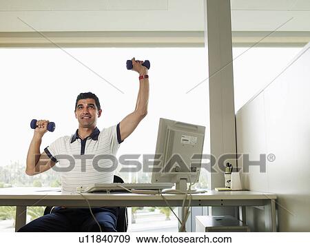 Stock Photo - Man sitting at desk lifting weights, smiling. Fotosearch