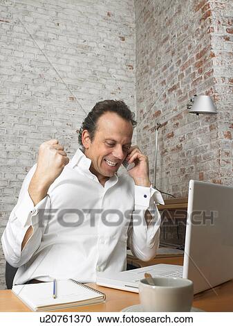 Stock Image - Mature businessman using phone in office, clenching fist, smiling. Fotosearch