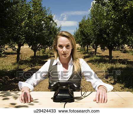 Stock Image - Woman sitting at desk behind old telephone in orchard.. Fotosearch