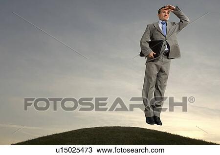 Stock Image - Businessman hovering above hill looking at horizon. Fotosearch