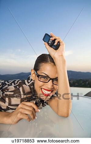 Stock Image - Woman leaning on table holding cellular phone smiling outdoors.. Fotosearch