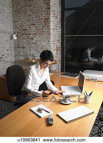 Stock Image - Businesswoman sitting at desk using mobile phone, night. Fotosearch