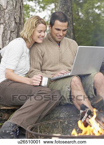 Stock Image - Couple by a fire pit with a laptop smiling.. Fotosearch