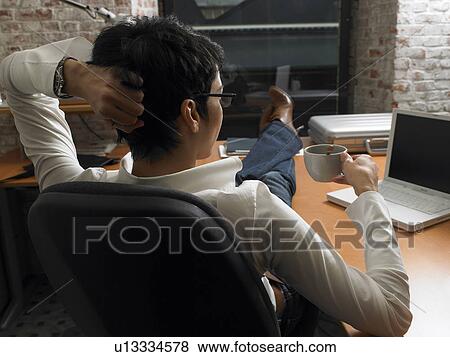 Stock Photo - Rear view of businesswoman having coffee with feet up on desk at dusk. Brussels, Belgium.. Fotosearch
