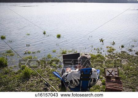 Stock Image - Senior man fishing by river, using laptop, rear view. Fotosearch
