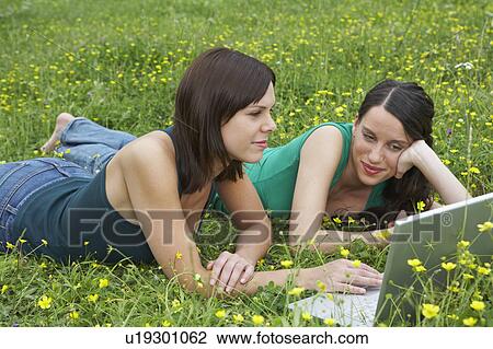 Stock Image - Two young women lying in meadow looking with laptop.. Fotosearch