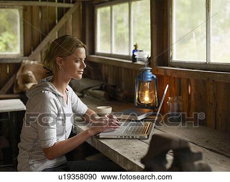 Stock Image - Woman with laptop and mug sitting at table in cabin.. Fotosearch