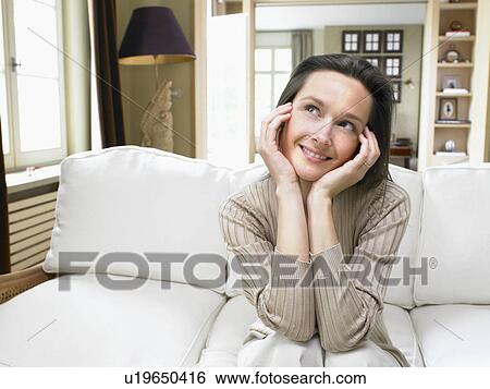 Stock Photograph - Woman sitting on sofa in living room smiling.. Fotosearch