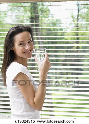 Stock Photography - Woman standing by a window holding a glass of water smiling.. Fotosearch