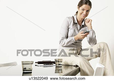 Stock Photo - Young business woman seated on desk sending text message.. Fotosearch