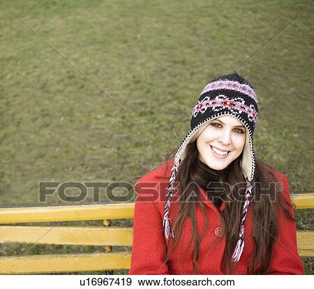 Stock Photo - Young woman wearing hat sitting on bench, smiling, portrait. Fotosearch