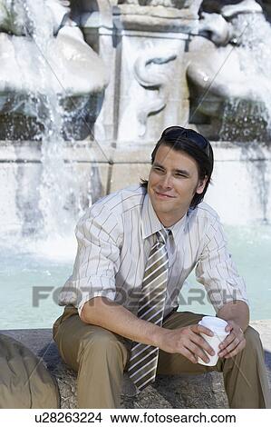 Picture - Businessman sitting on edge of fountain with coffee to go smiling.. Fotosearch