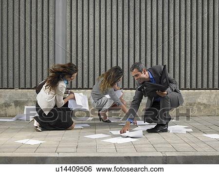 Stock Photograph - Two businesswomen and one businessman trying to recover papers blowing about pavement, Alicante, Spain,. Fotosearch