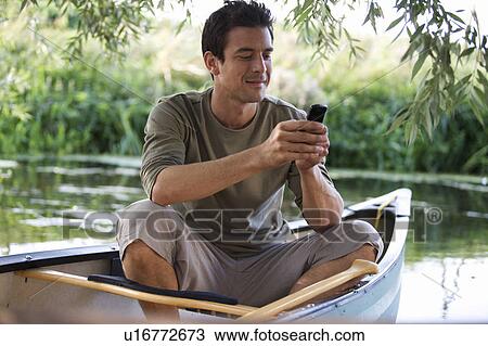 Stock Image - Young man sitting in a boat talking on a mobile phone. Fotosearch