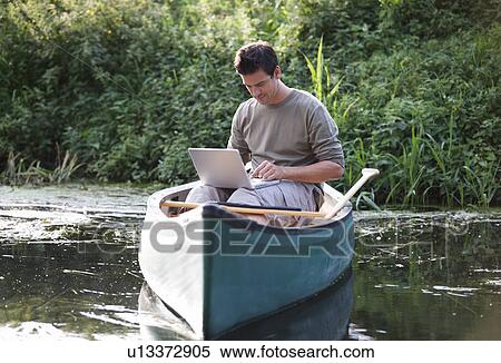 Stock Photography - Young man sitting in a boat working on a laptop. Fotosearch