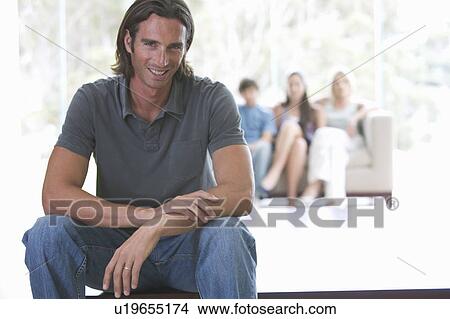 Picture - A father sitting on a coffee table with his family sitting in a sofa. Fotosearch