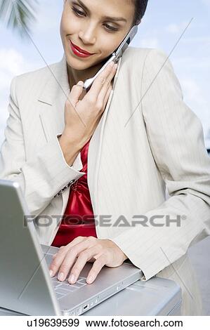 Stock Photo - A businesswoman talking on a mobile phone using a laptop computer. Fotosearch