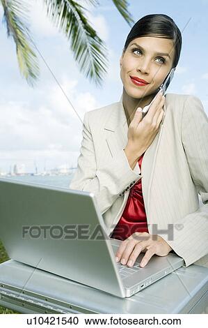 Stock Image - A businesswoman talking on a mobile phone using a laptop computer. Fotosearch